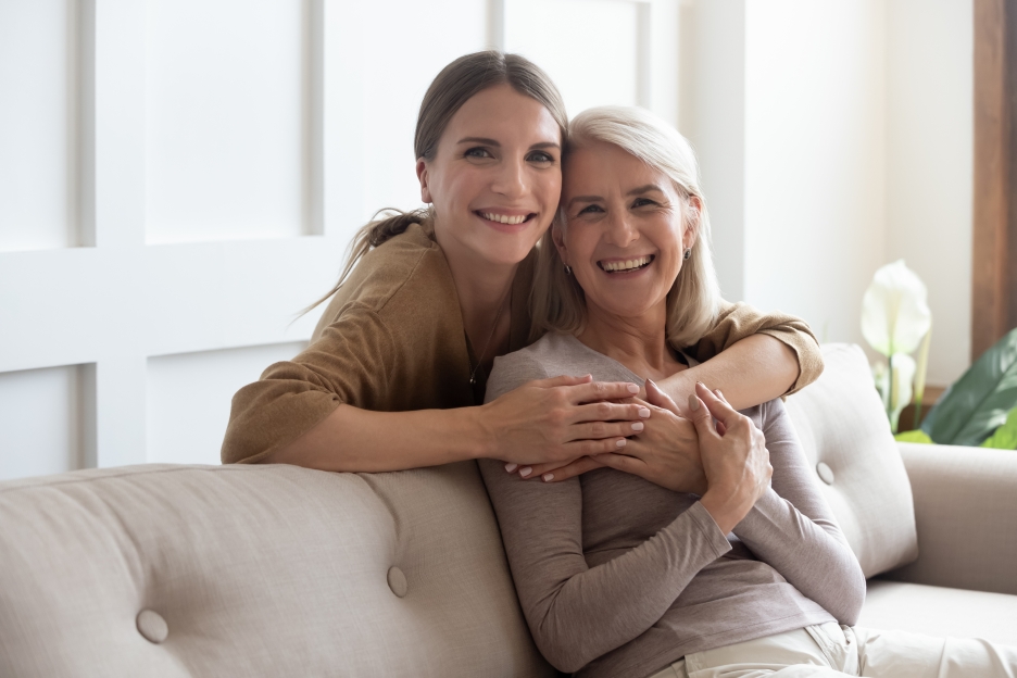 Smiling woman and older woman embracing on a sofa, representing family connections and wellness, relevant to Front Door Medspa's aesthetic and health services.